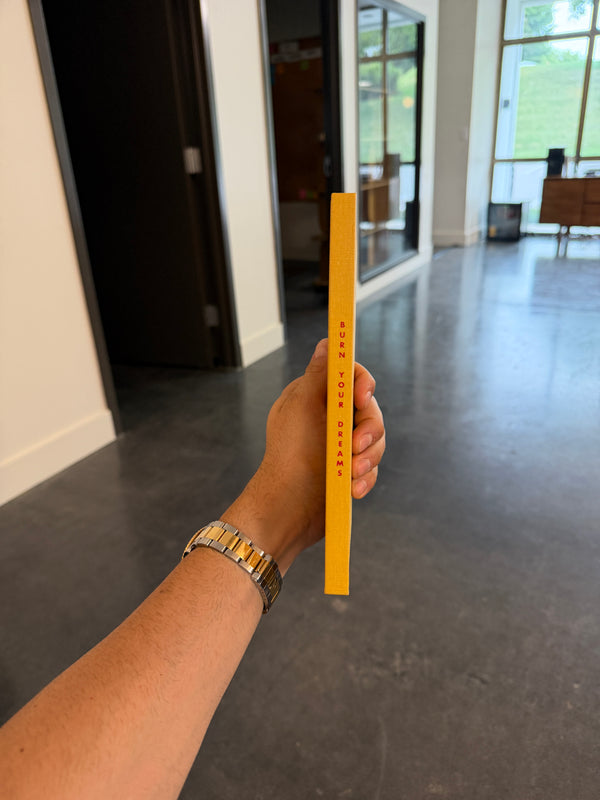 A hand holding a slim yellow book or card with vertical red text that reads 'burn your dreams' in an indoor hallway with a concrete floor and large windows in the background.