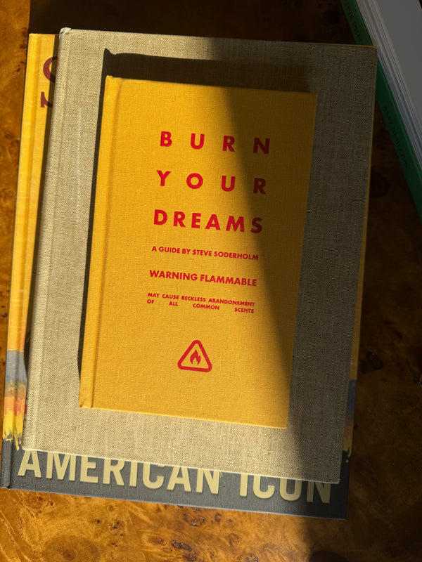 A book titled 'Burn Your Dreams' by Steve Soderholm, featuring a bright yellow cover with red text and a warning flame icon, resting on top of a larger gray book on a wooden surface.