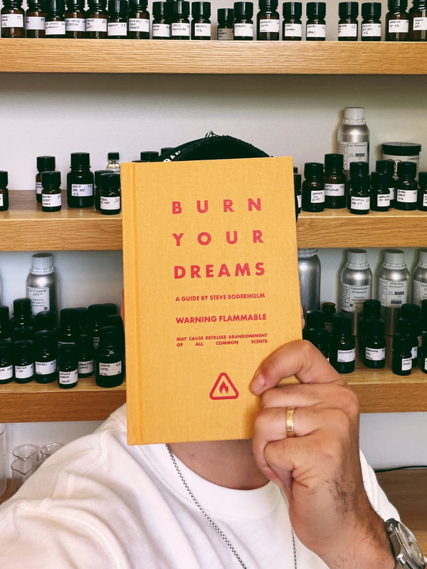 A person holding a yellow book titled 'BURN YOUR DREAMS' in front of their face, with shelves of small bottles in the background.