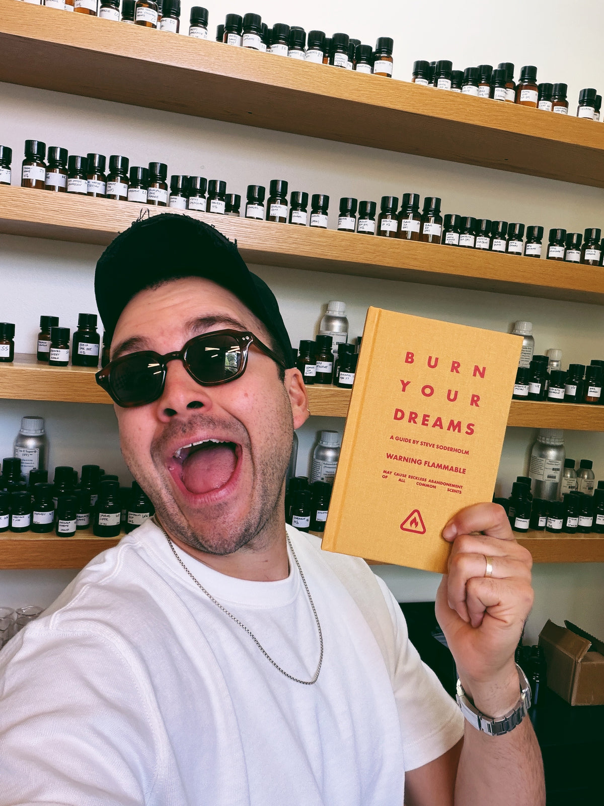 A man wearing a black cap, sunglasses, and a white t-shirt holds up a yellow book titled 'BURN YOUR DREAMS' in front of shelves filled with small bottles.