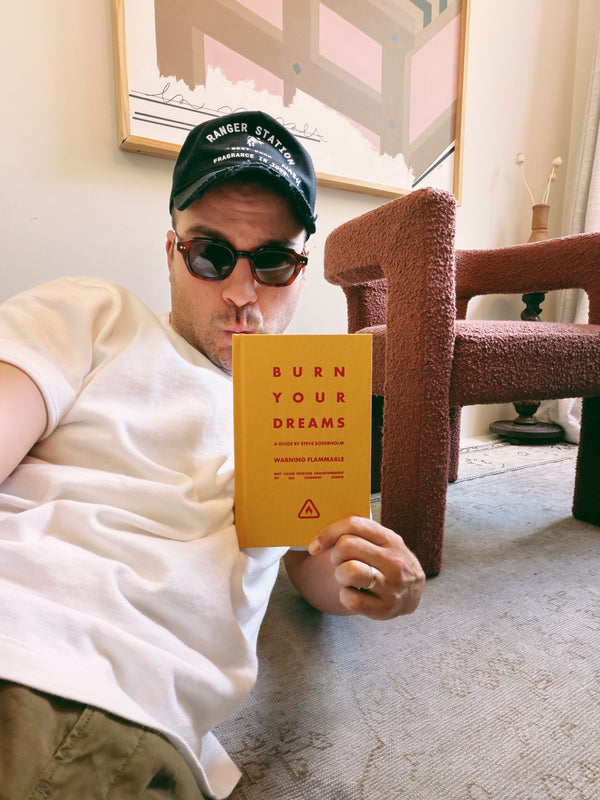 A man wearing a black cap and sunglasses holds up a yellow book titled 'BURN YOUR DREAMS' in a cozy indoor setting with a textured brown armchair and framed art in the background.
